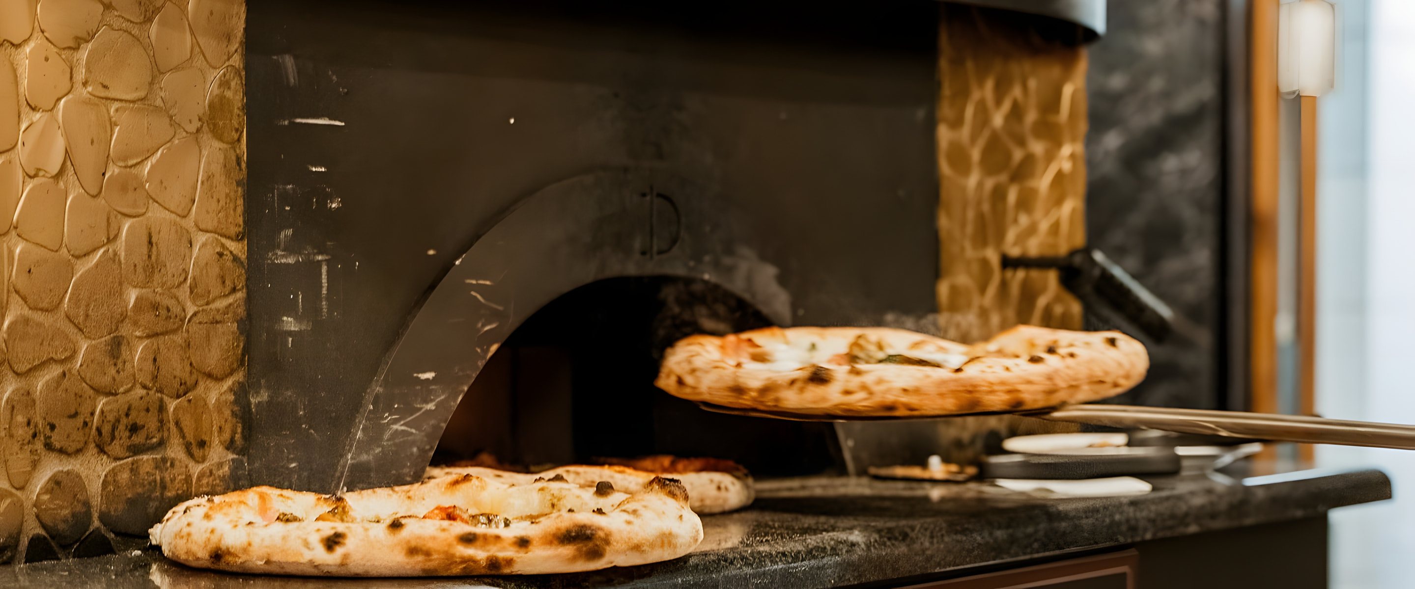 Goldener Mosaik-Kuppelofen der Pizzeria Capri: Eine Pizza kommt auf der Schaufel aus dem Ofen, zwei fertige Pizzen liegen davor