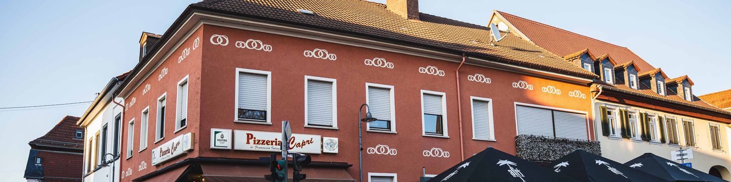 Die rote Fassade der Pizzeria Capri am Marktplatz im Abendlicht, mit dem Schild „Pizzeria Capri“ und Sonnenschirmen